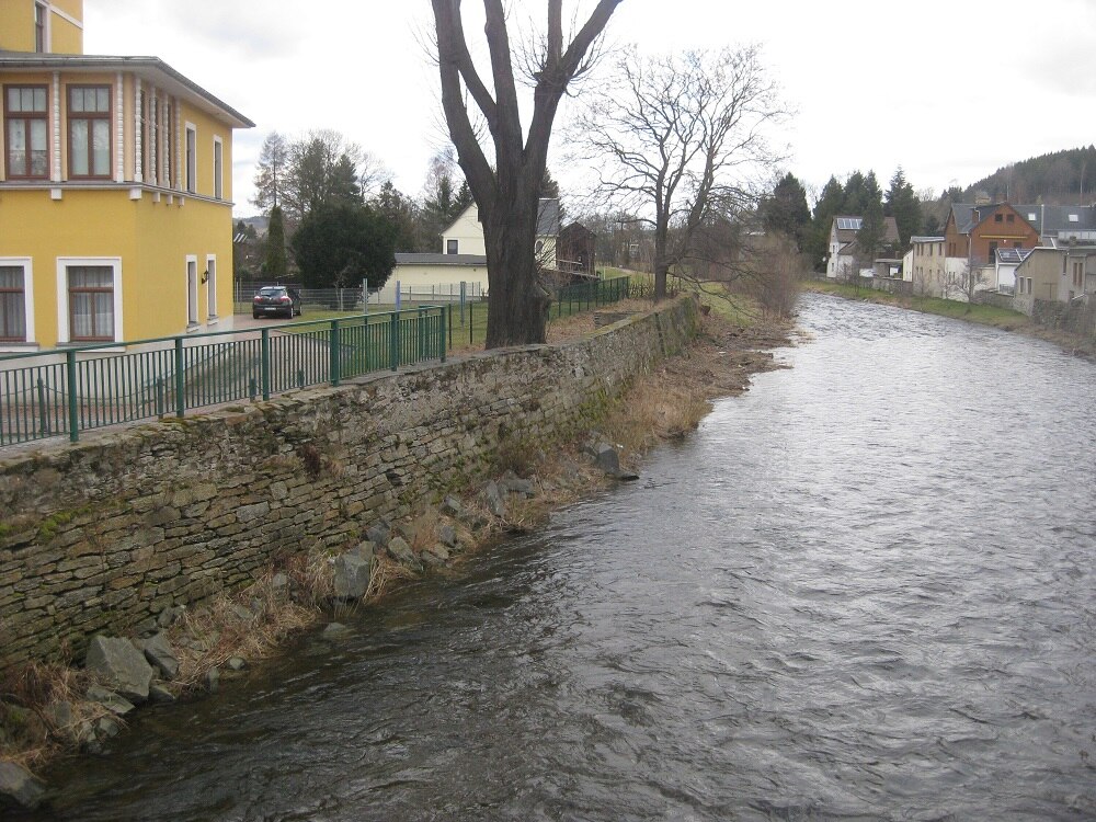 Ein Fluss, am linken Ufer eine Mauer darauf ein grünes Geländer und dahinter ein gelbes Haus