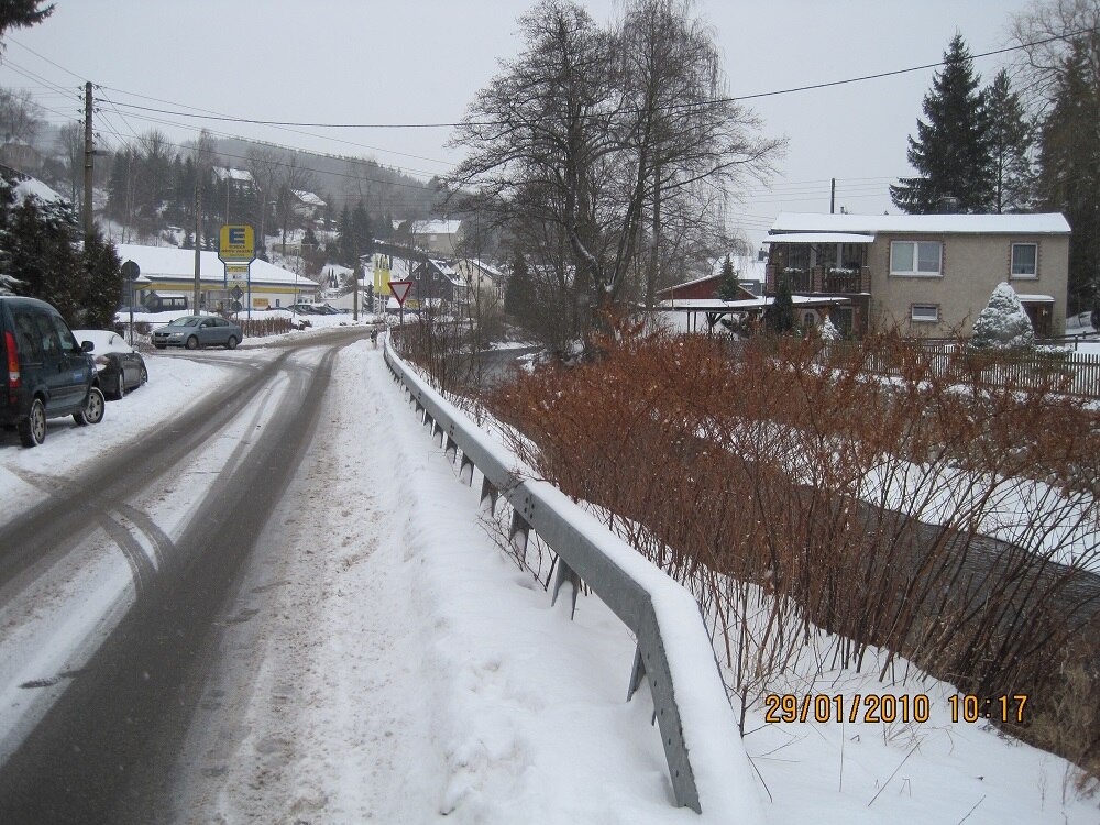 verschneite Straße mit Leitplanke, dahinter Häuser