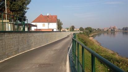 Blick auf asphaltierte Straße, rechts ein grünes Geländer, dahinter ein Fluss, links eine Mauer dahinter ein Wohnhaus