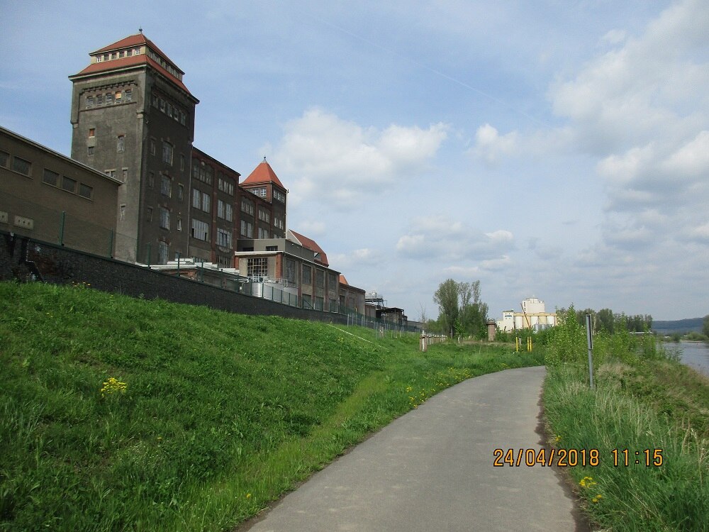 Links im Bild eine große alte Fabrik, davor ein mit Gras begrünter Hang, vor dem ein asphaltierter Weg verläuft, rechts daneben Wiese und ein Fluss