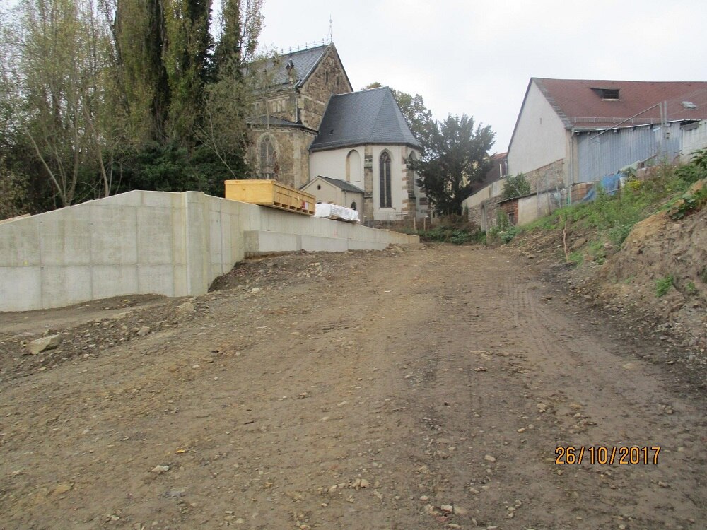 Blick einen Schotterweg hinauf, links davon eine Betonmauer, dahinter eine Kirche