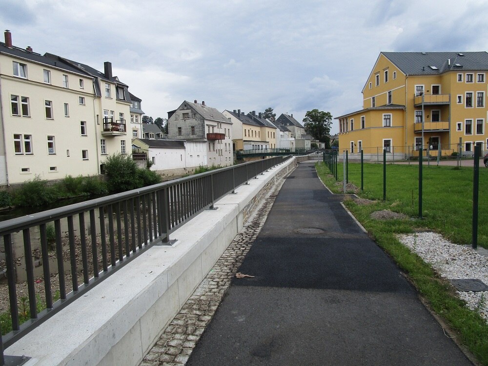 asphaltierter Weg mit niedriger Mauer und Geländer links, im Hintergrund Häuser