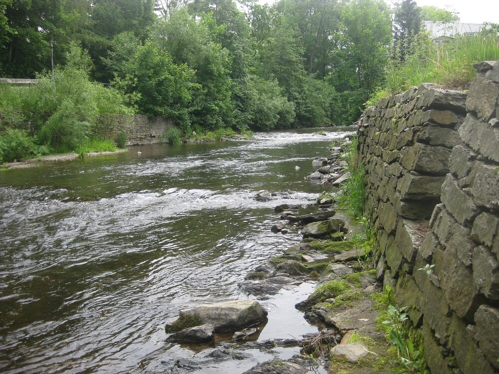 Fluss mit Bäumen am linken Ufer und einer Mauer am rechten Ufer