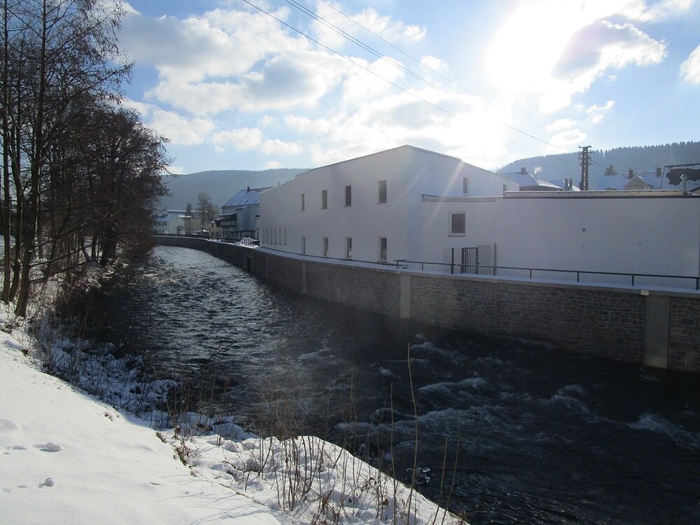Blick von einem schneebedeckten Ufer auf einen Fluss an dessen anderem Ufer eine Mauer mit einem weißen Gebäude dahinter zu sehen ist