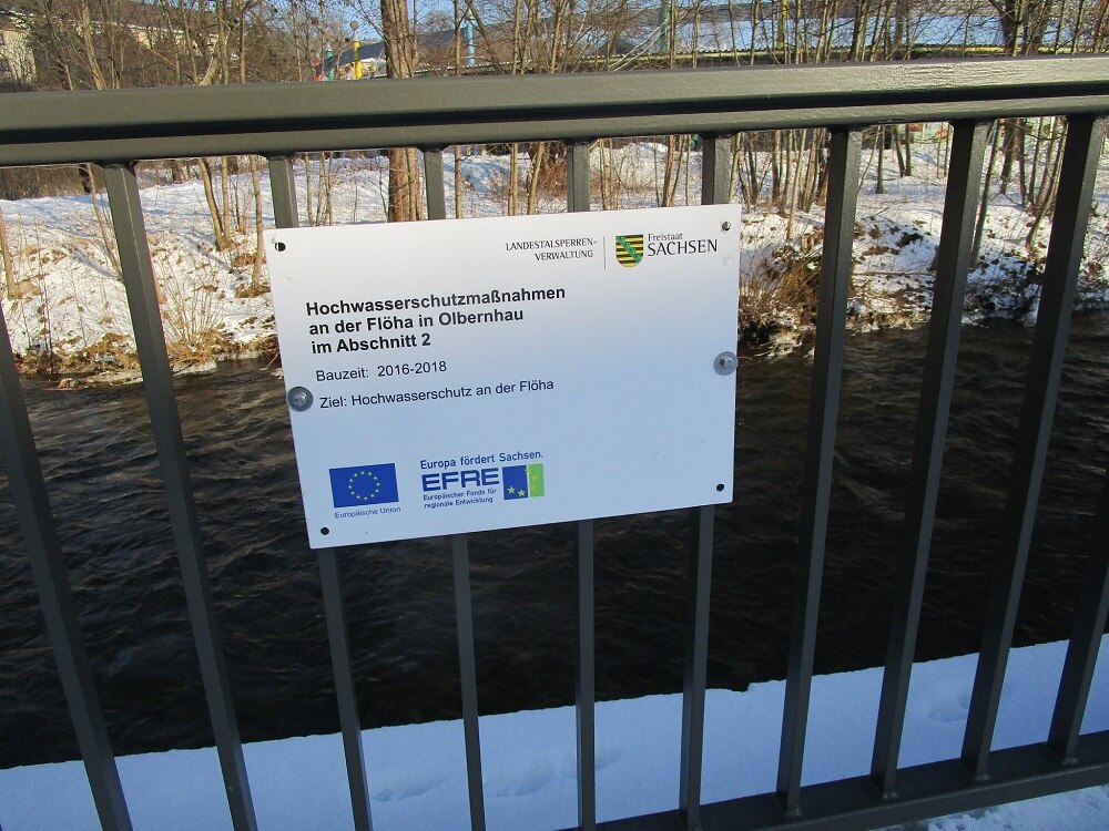 Ein kleines Infoschild an einem Geländer neben einem Fluss