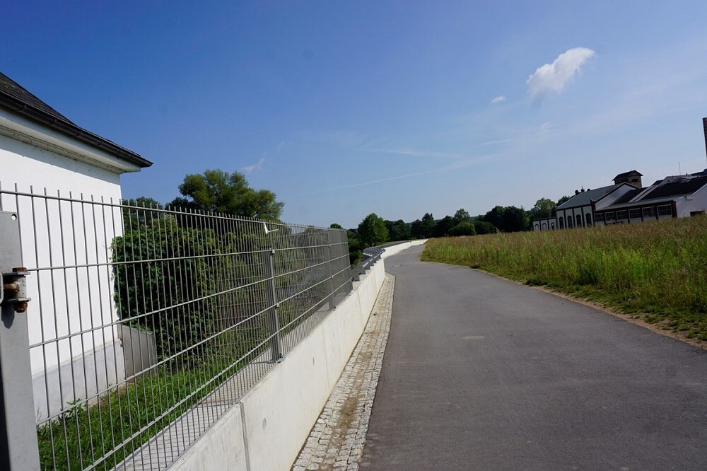 asphaltierter Weg, links niedrige Mauer mit Zaun darauf, dahinter ein Haus, rechts vom Weg Wiese