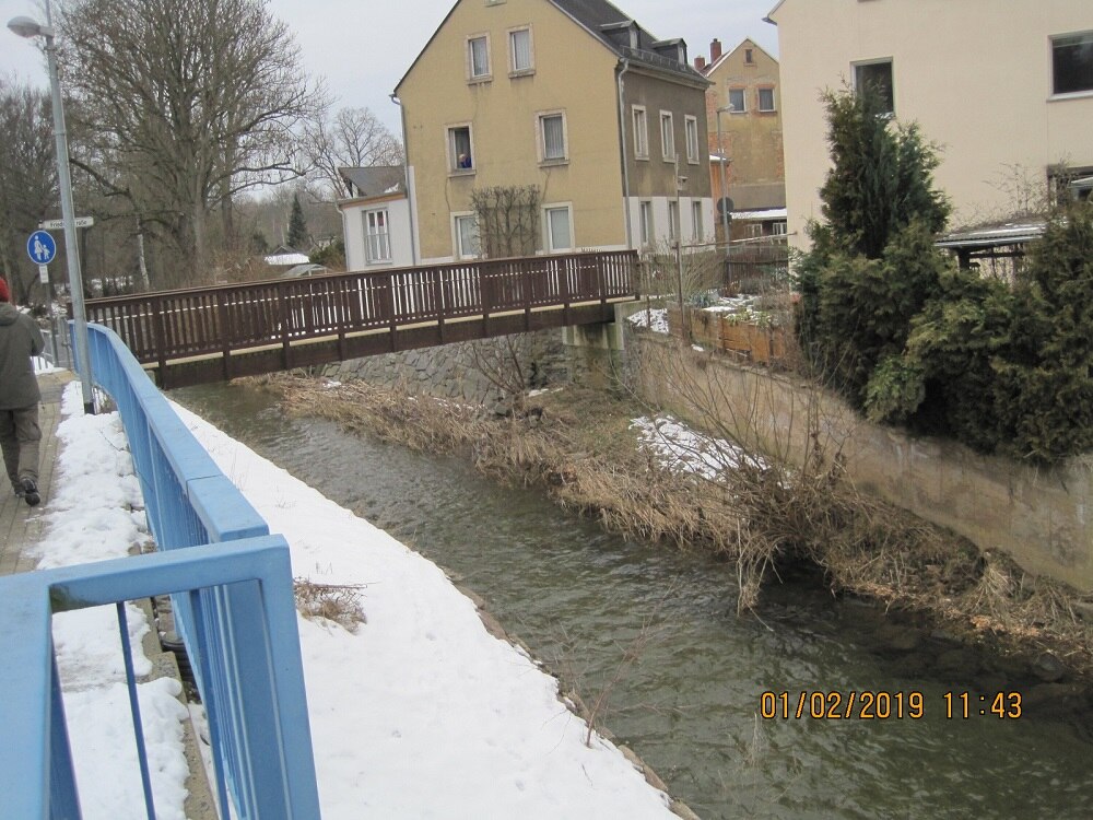 braune Holzbrücke über einen Fluss, dahinter Häuser