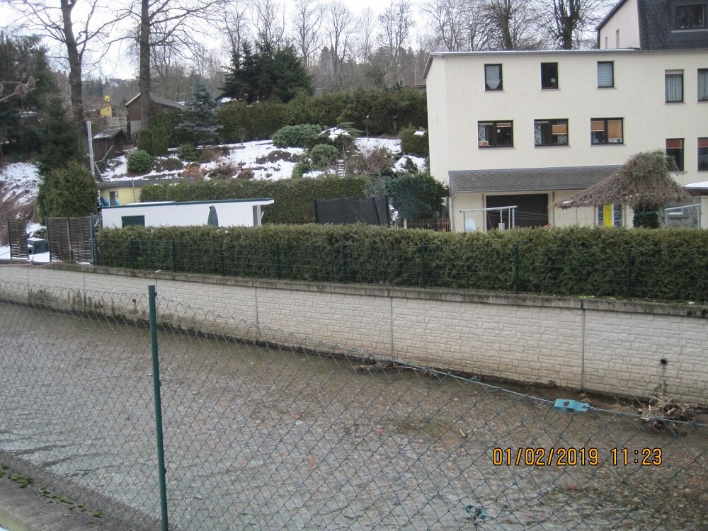 Maschendrahtzaun, dahinter Fluss mit Ufermauer, dahinter Häuser