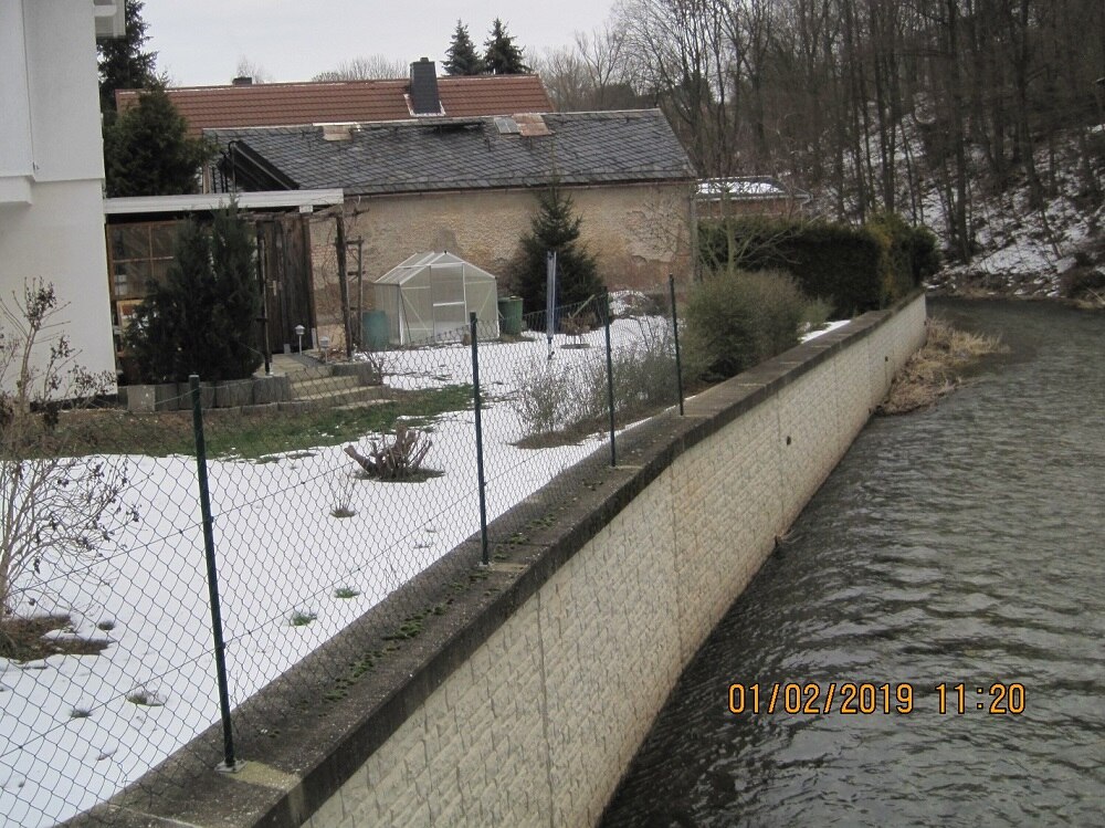 rechts ein Fluss, links davon Ufermauer und angrenzende Grundstücke