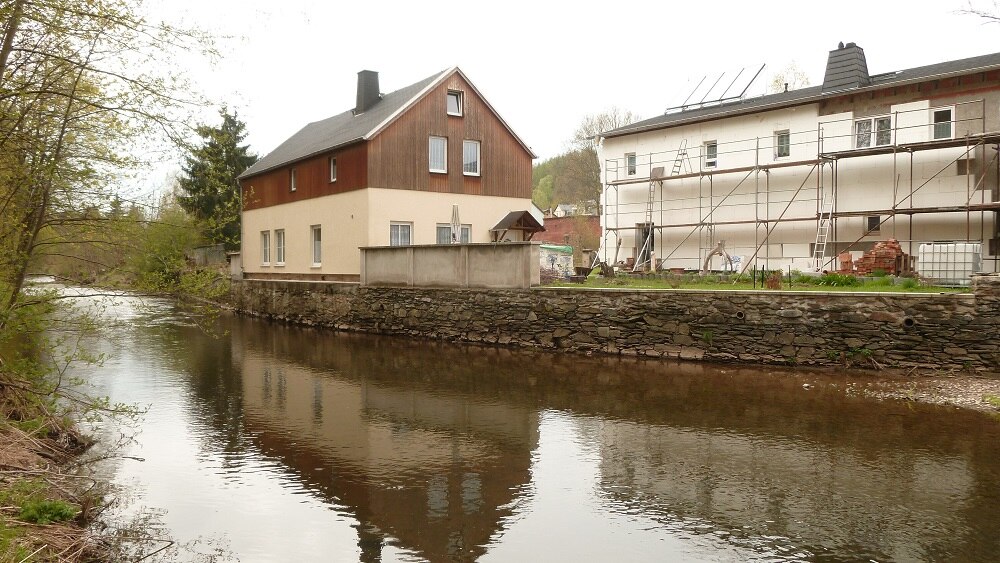 Im Vordergrund ein Fluss, am gegenüberliegenden Ufer stehen Häuser