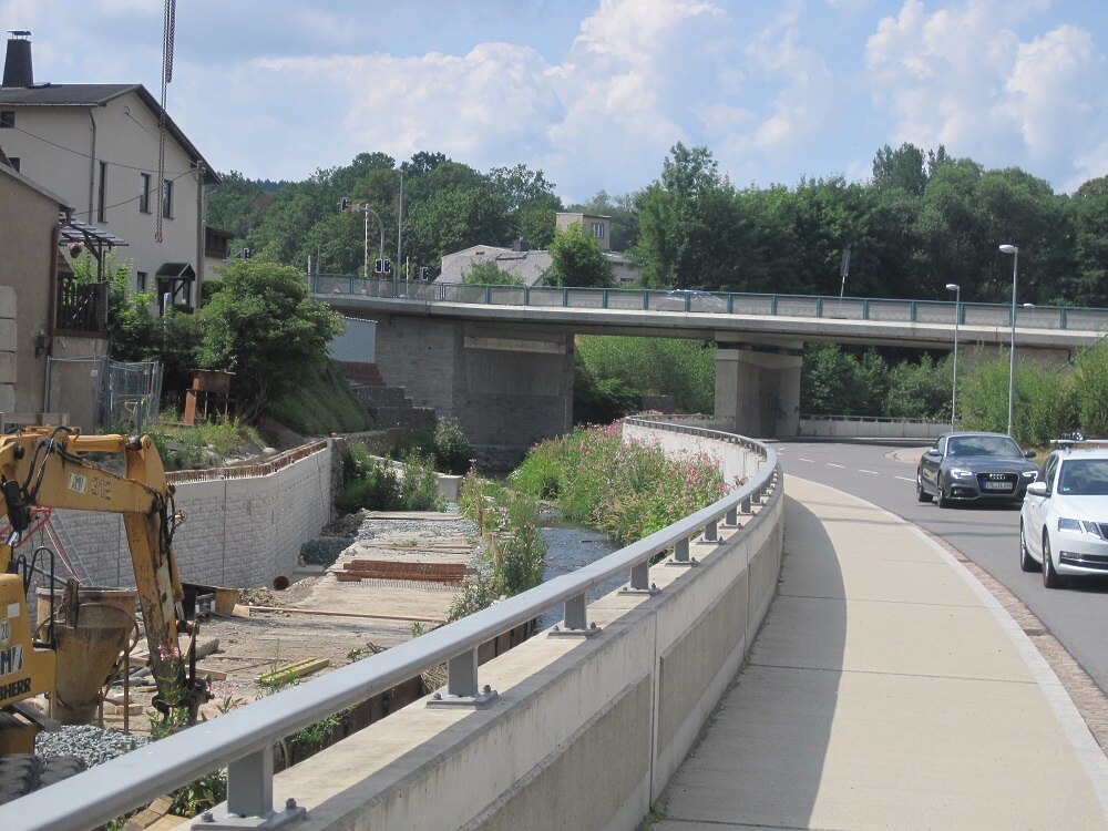 Blick auf Straße mit niedriger Mauer mit Geländer, im Hintergrund eine Brücke, links Bagger und Häuser