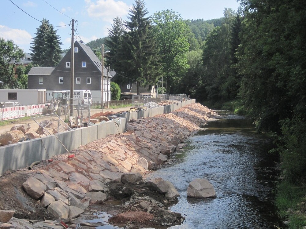 Blick auf Flussufer im Bau, im Hintergrund ein Haus und Wald