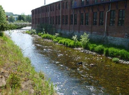 Blick von begrüntem Ufer auf die andere Uferseite eines Flusses, an die direkt ein langes Backsteingebäude anschließt