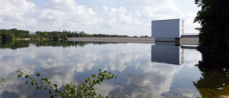 Wolken spiegeln sich in der Wasseroberfläche, in der der Entnahmeturm der Talsperre Bautzen steht. Im Hintergrund die Staumauer.
