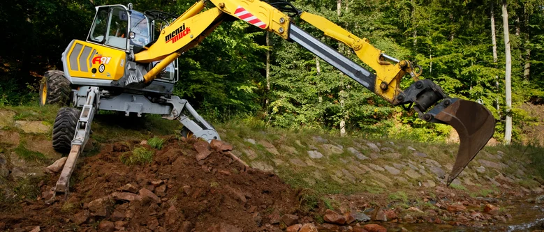 ein gelber Spezialbagger mit zusätzlichen Stützen steht auf einer Böschung aus großen Steinen. Im Hintergrund grüner Wald.