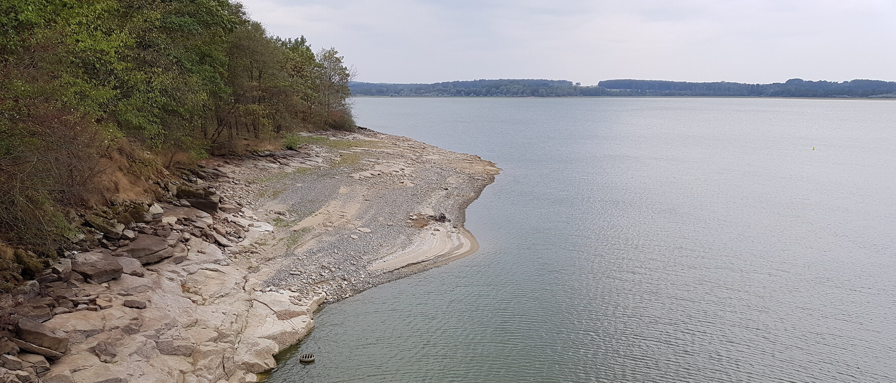 See mit breitem Steinufer links und Wald