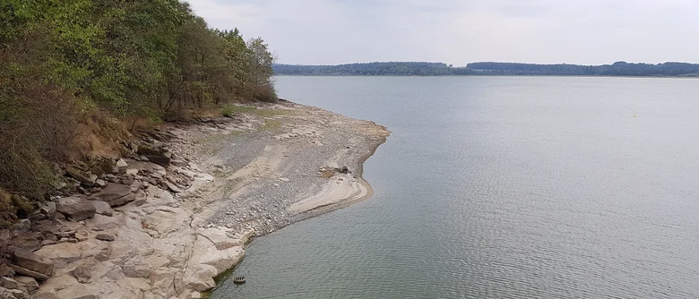 See mit breitem Steinufer links und Wald