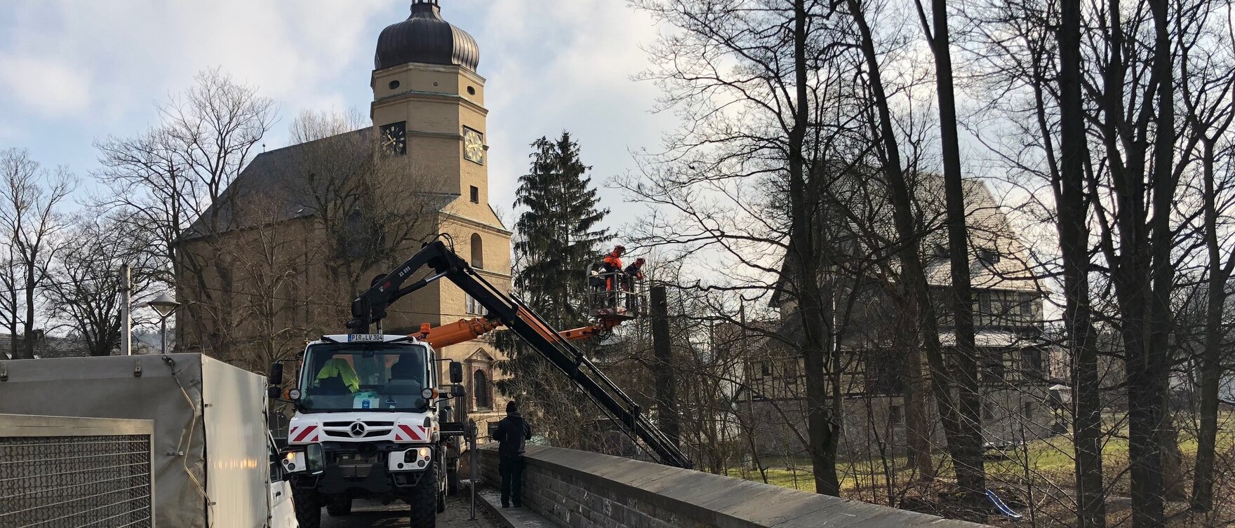 Spezialfahrzeug mit Hebebühnen auf denen zwei Arbeiter vor Bäumen stehen. Im Hintergrund eine Kirche und ein Fachwerkhaus.
