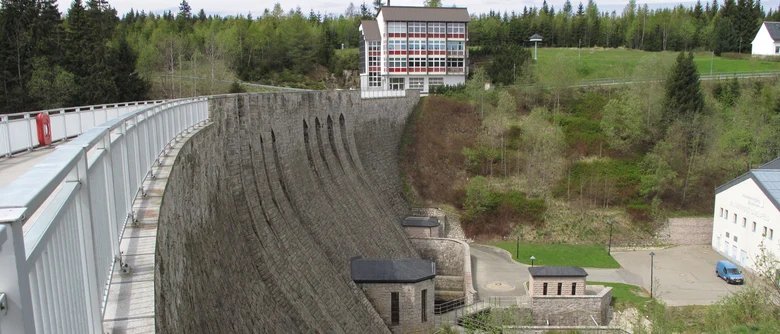 Staumauer mit weiß-rotem Gebäude am Ende umgeben von Wald