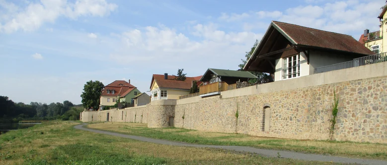 Wiese neben einem Fluss mit Weg und angrenzender Mauer mit Häusern