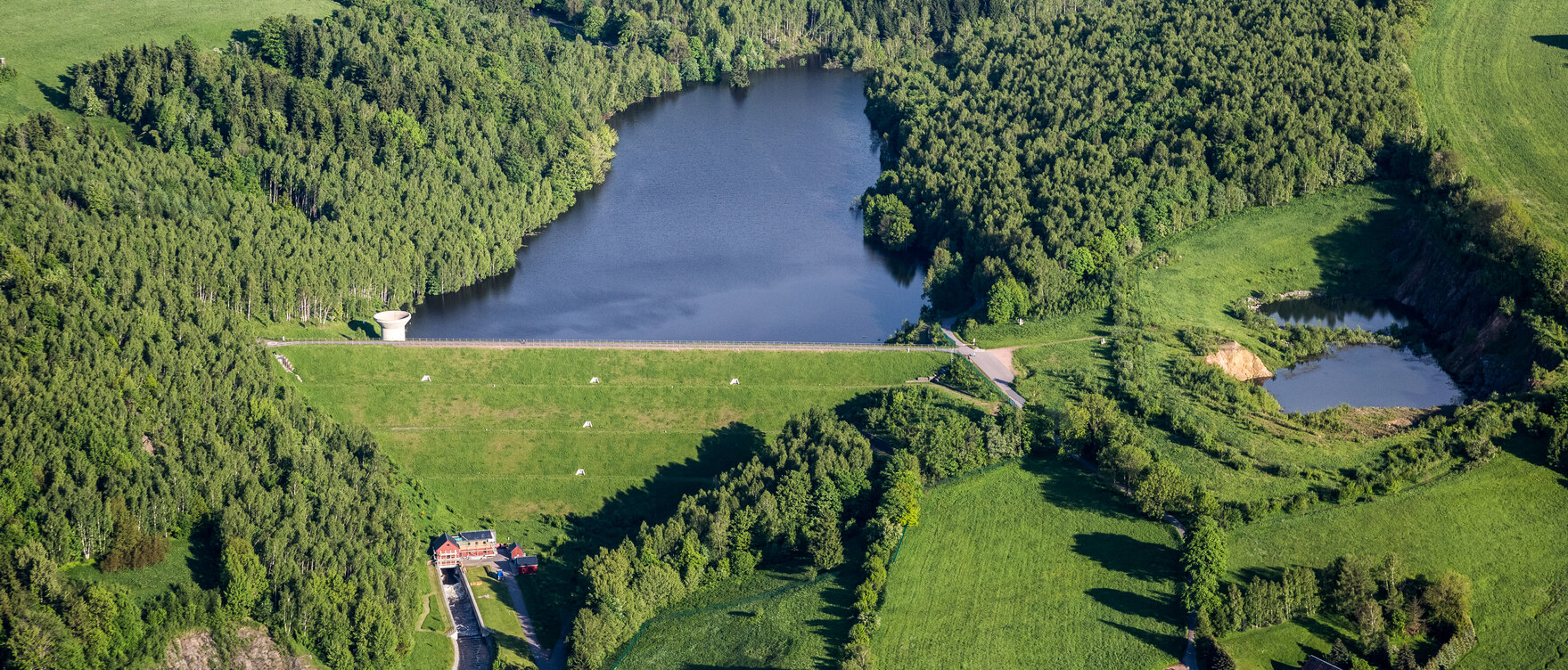 Blick vom mit Gras bewachsenen Ufer auf das Wasser im Hochwasserrückhaltebecken Lauenstein. Im Vordergrund steht ein Trichter aus Beton. Der See ist umgeben von Wald.