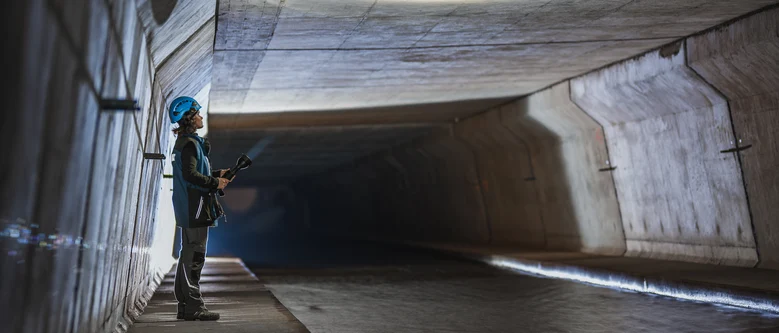 Eine Frau mit blauem Helm auf dem Kopf und Taschenlampe in der Hand steht in einem dunklen Tunnel und schaut an die Decke
