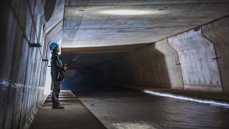 Eine Frau mit blauem Helm auf dem Kopf und Taschenlampe in der Hand steht in einem dunklen Tunnel und schaut an die Decke