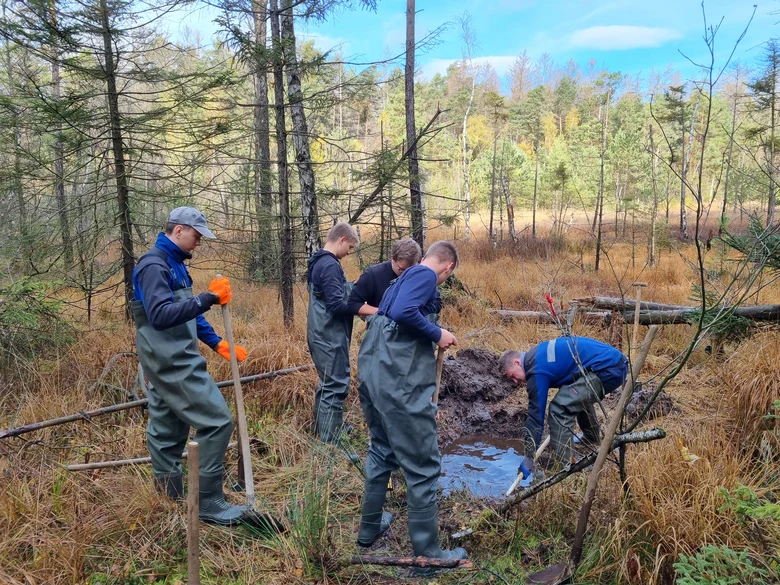 fünf Auszubildende stehen im Wald um ein Loch mit Wasser. Einer schaufelt.italisierung