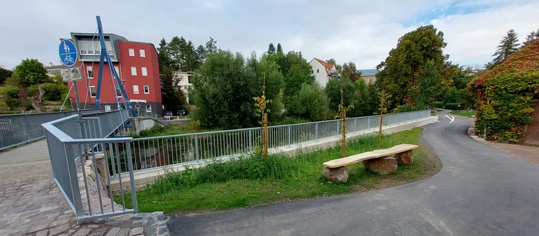 Blick auf einen Fluss mit neu errichteter Spundwand und einem Fußgängerweg daneben, im Vordergrund eine Bank aus Holz und Stein, im Hintergrund Wohnhäuser und Bäume
