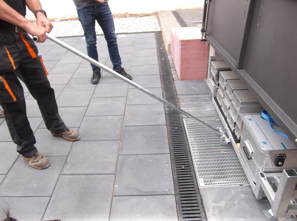 graue Gehwegplatten mit Regenablauf und Balken und eine Kiste aus Metall unter einem dunkelgrauen Tor