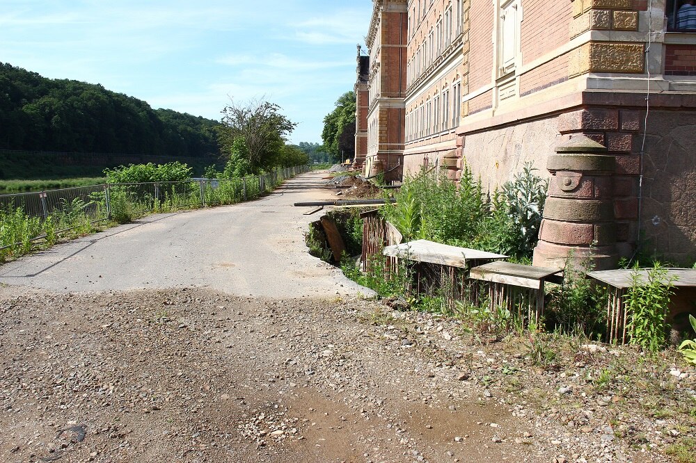 Bick auf einen Schotterweg der in einen asphaltierten Weg über geht entlang eines alten Gebäudes, im Hintergrund Wald