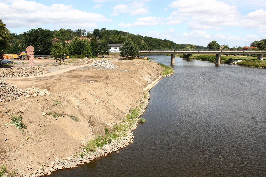 Ein Fluss mit Schotterböschung, im Hintergrund eine Brücke