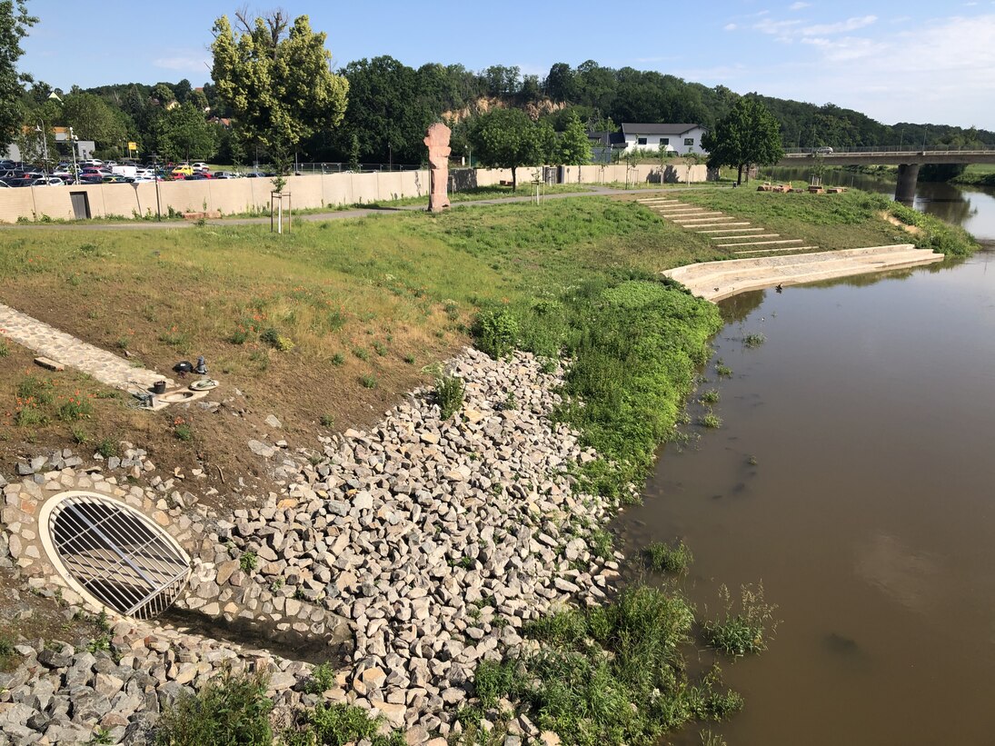 eine begrünte Böschung mit gröberen Steinen am Flussufer und einem vergitterten Auslass, eine Treppe am Ufer und im Hintergrund eine Betonmauer vor einem Parkplatz mit Autos