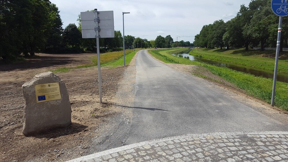 asphaltierte Straße parallel zu einem Fluss, links ein Findling mit einem Metallschild