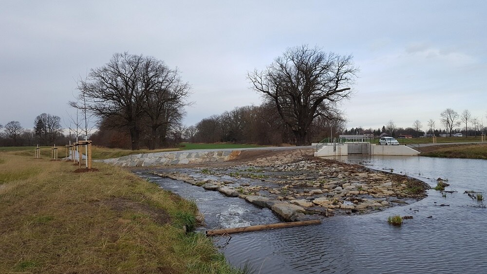 Blick auf das Wehr aus der Ferne zwischen Wiese und Bäumen