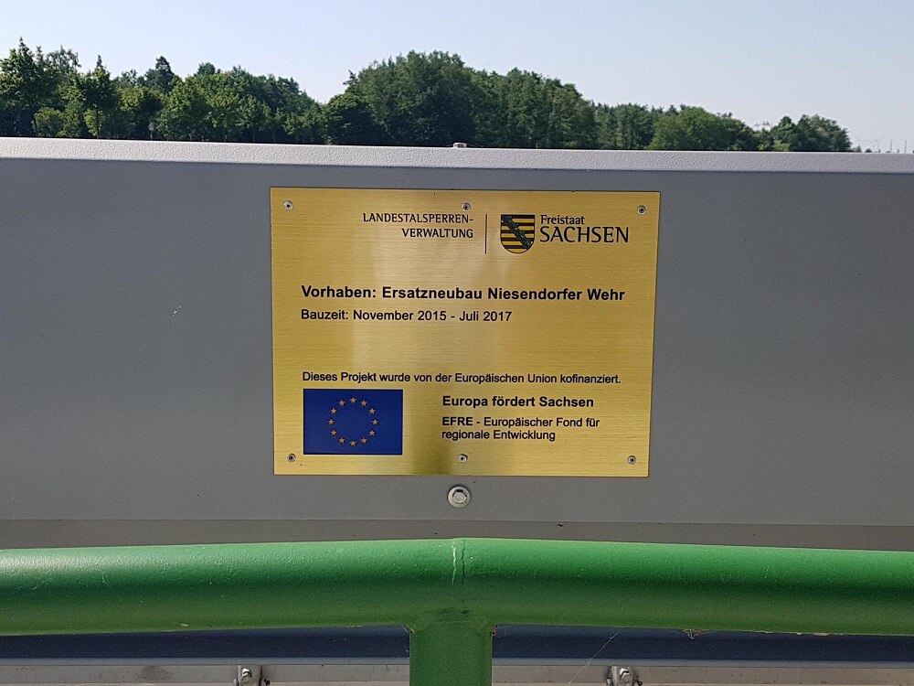 Goldenes Metallschild mit Informationen zur EU-Förderung auf einer Mauer, davor ein grünes Geländer
