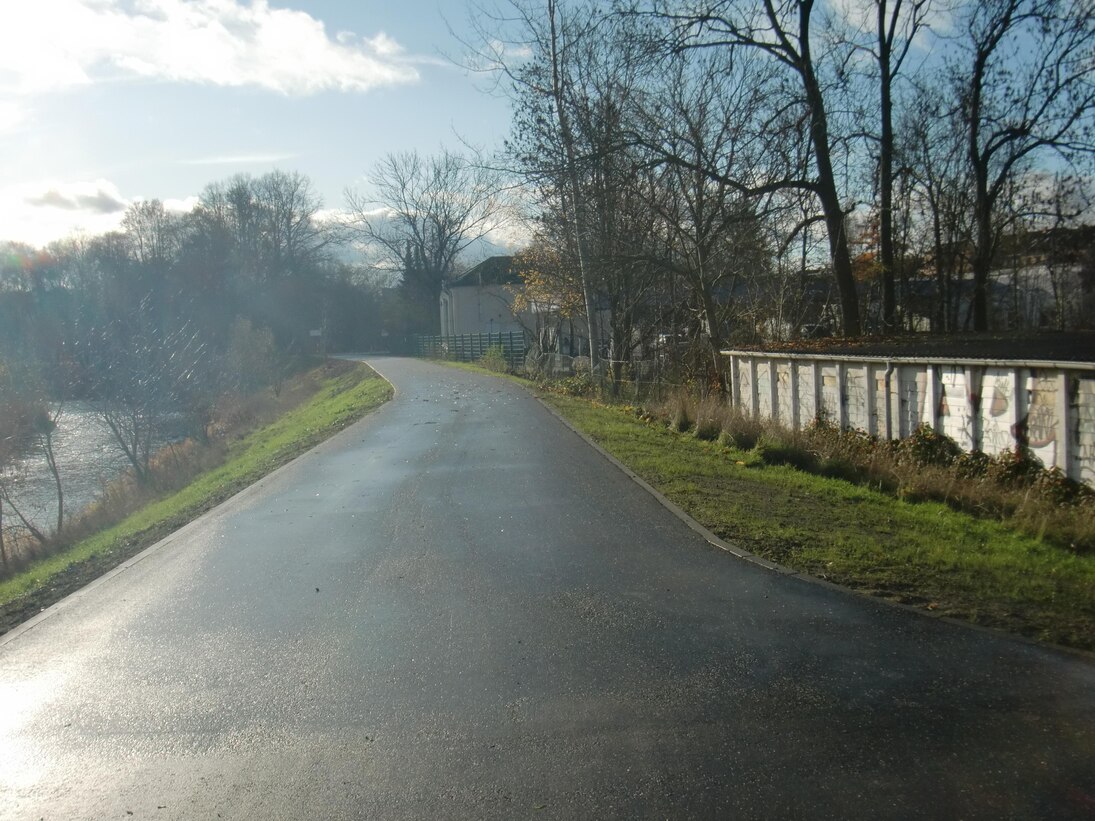 Blick auf eine asphaltierte Straße, rechts davon ein Grünstreifen, dahinter Gebäude. Links vom Weg eine begrünte Böschung hin zu einem Fluss