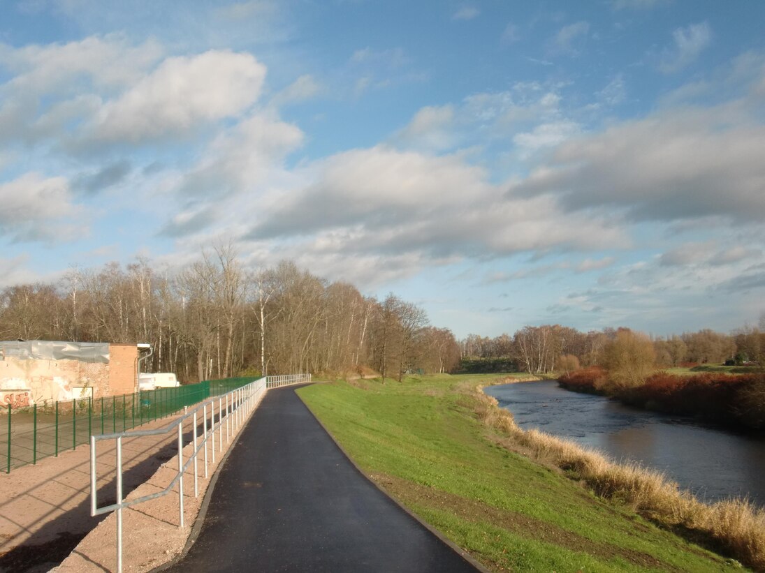 Blick auf einen asphaltierten Weg mit einem Metallgeländer auf der linken Seite und einer begrünten Böschung und einem Fluss auf der rechten Seite