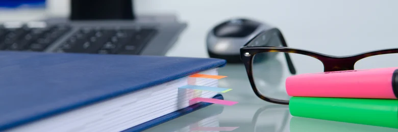 Notizbuch liegt neben Textmarkern in pink und grün vor einer Brille. Im Hintergund befindet sich eine Tastatur und eine dazugehörige Maus
