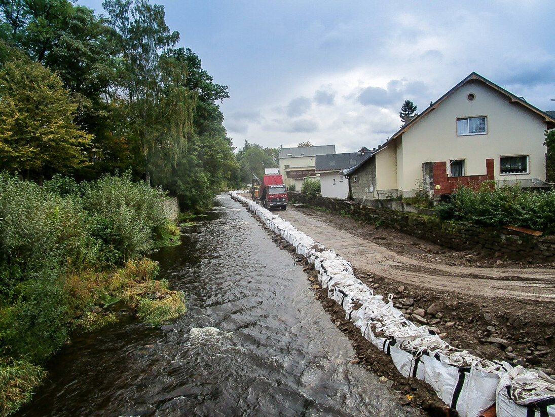 Blick auf den gleichen Fluss, der mit Sandsäcken abgegrenzt ist daneben eine Baustraße und dann die Häuser