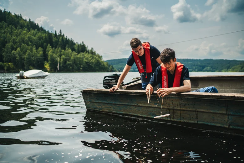 zwei junge Männer sitzen ini einem Boot auf dem Wasser und haben Messgeräte in der Hand. Im Hintergrund Wälder und ein paar Wolken am Himmel