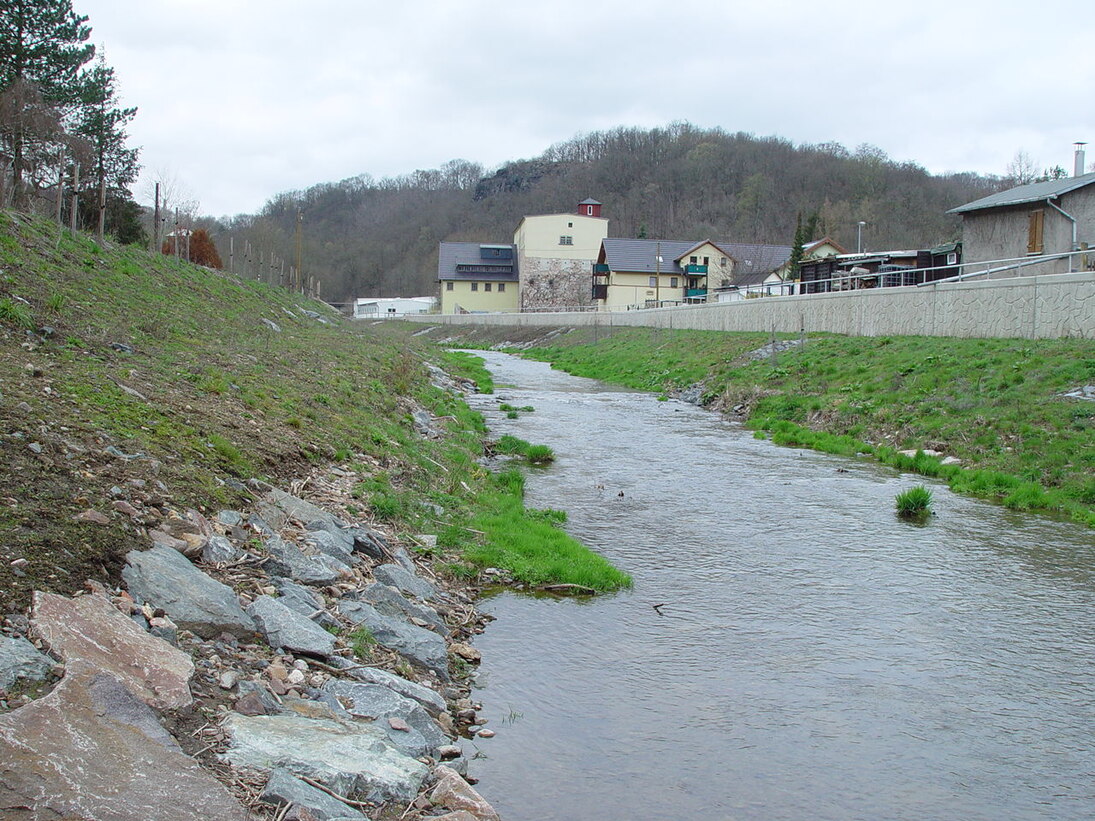Blick auf einen Fluss an dessen linkem Ufer Natursteine und Wiese zu sehen ist und entlang des rechten Ufers eine Mauer, hinter der sich Häuser befinden