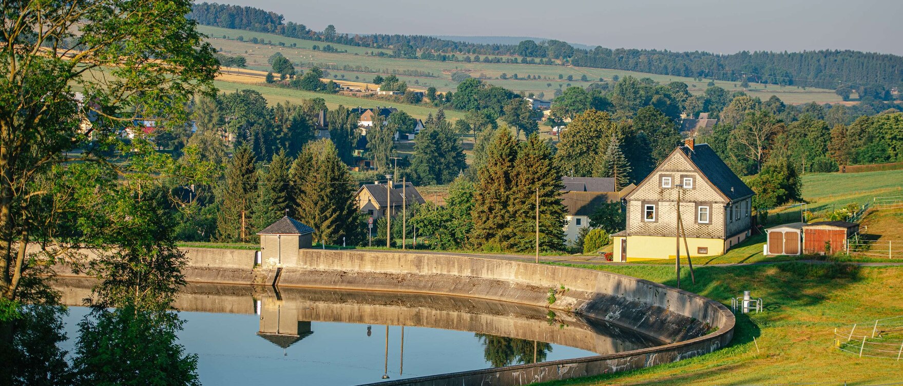 Blick auf einen Teich, der von einer Mauer umgeben ist, dahinter Häuser, Bäume und Felder
