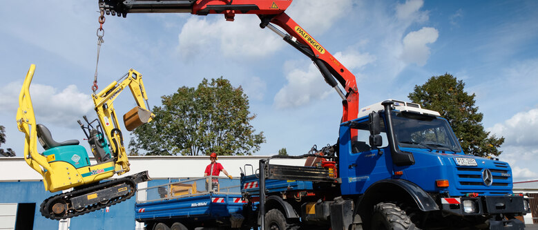 ein kleiner gelber Raupenbagger wird mit einem Kran auf den Hänger eines großen Arbeitsfahrzeuges geladen