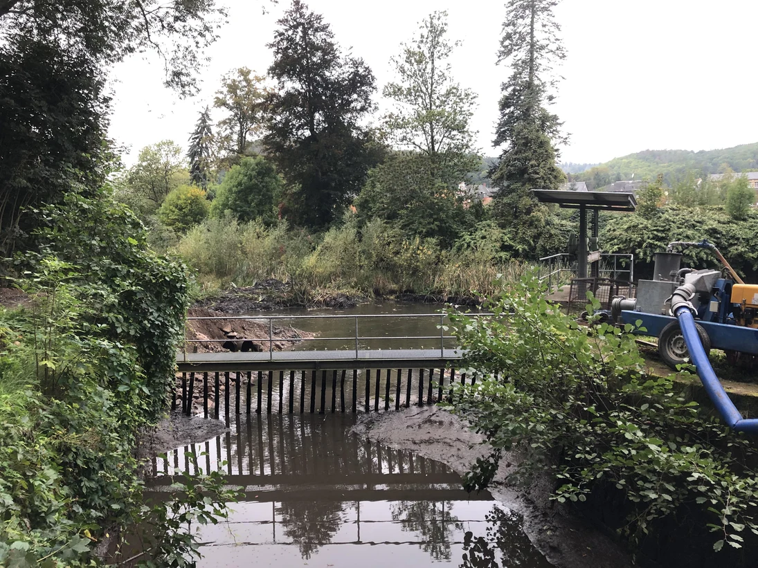 Flusslauf mit Brücke und Metallgitter, umgeben von dichtem Grün und Bäumen, mit einer blauen Rohrleitung rechts im Bild
