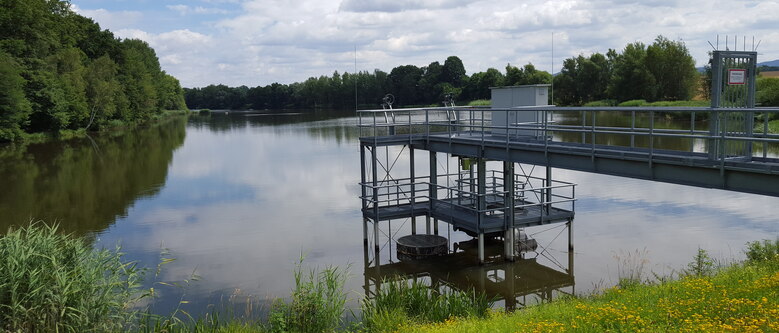 Blick vom Ufer auf einen von Wald umgebenen See in den ein Steg ragt