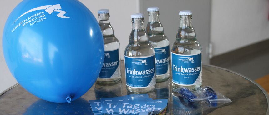 ein blauer Luftballon, drei Flaschen und Flyer liegen beziehungsweise stehen auf einem Tisch