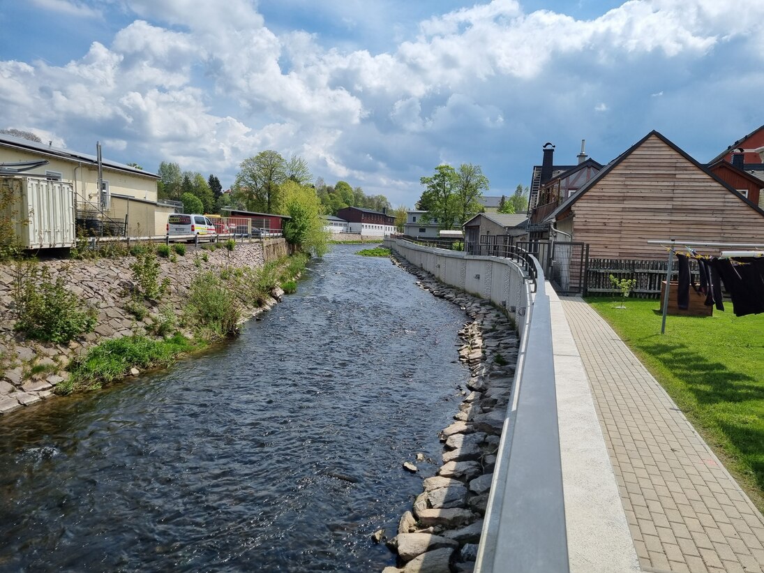 Blick auf Fluss mit Häusern rechts und links und einer neuen Hochwasserschutzmauer auf der rechten Seite darüber blauer Himmel mit Wolken