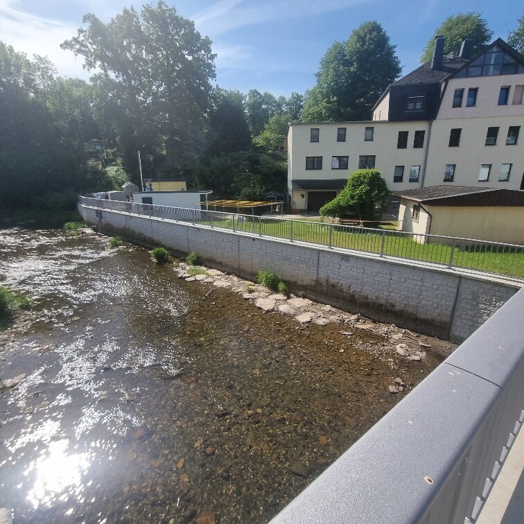 Blick auf Fluss mit Mauer rechts und Mehrfamilienhaus dahinter