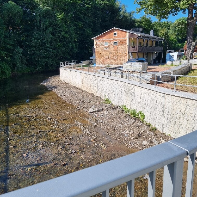 Blick von Brücke auf Fluss mit Bäumen im Hintergrund und einer Mauer rechts, dahinter ein Haus